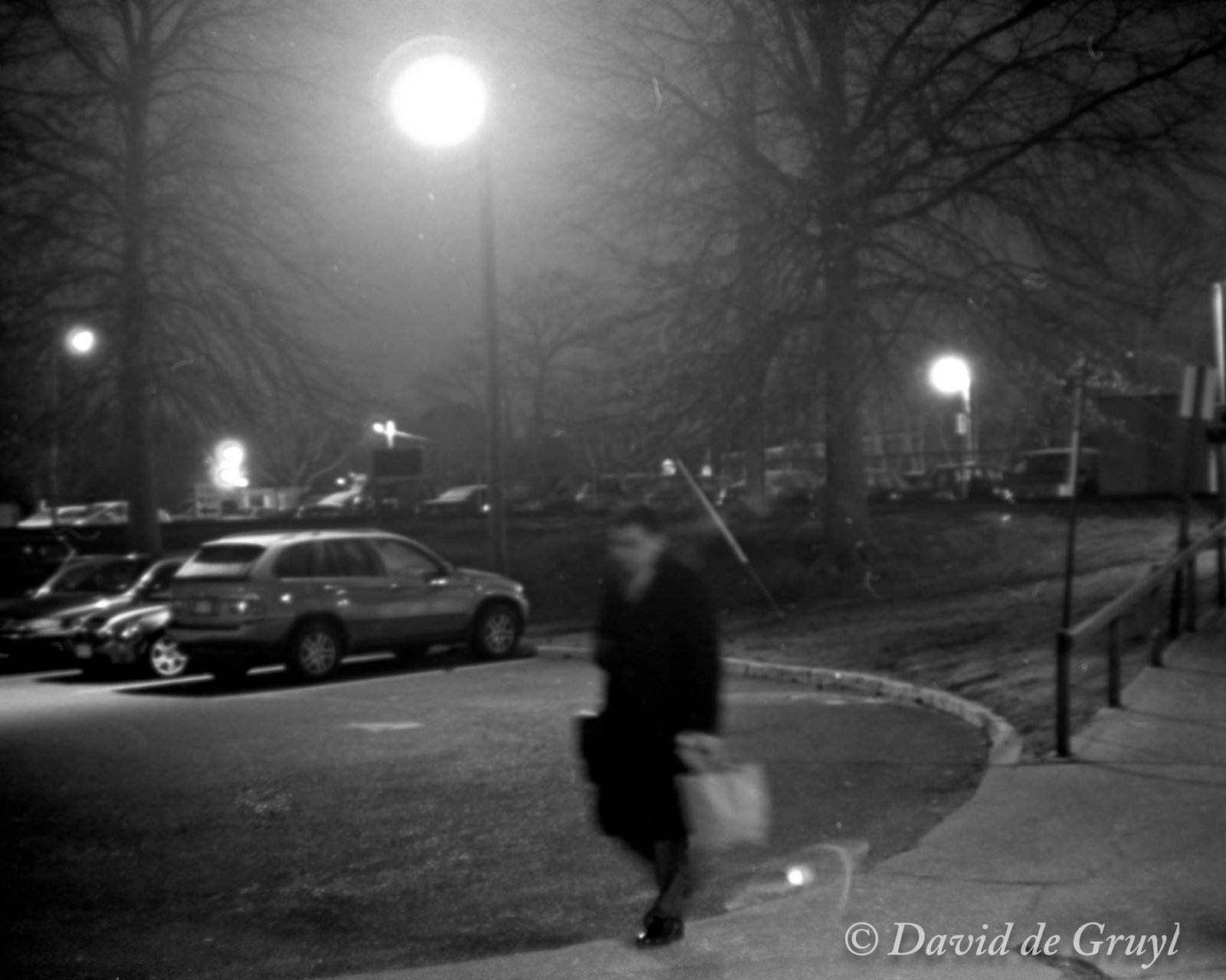 Black and white print of a person walking at night in a train station parking lot